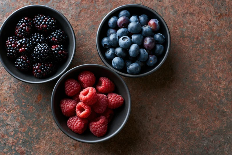 Three dark ceramic bowls filled with blackberries, blueberries and raspberries arranged on a weathered rust-brown surface