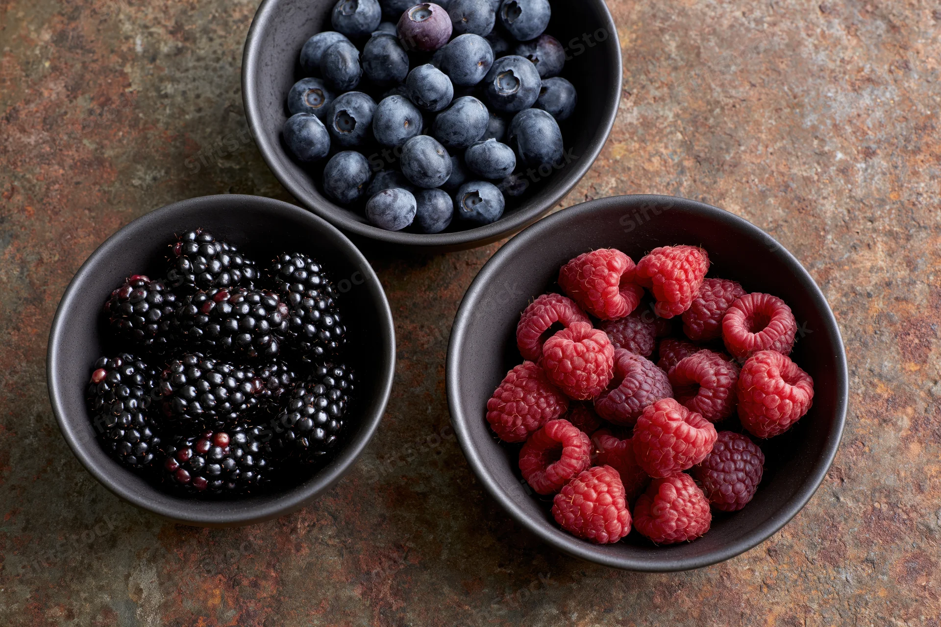 Three dark ceramic bowls each filled with blackberries, raspberries and blueberries arranged on a weathered rust-brown surface