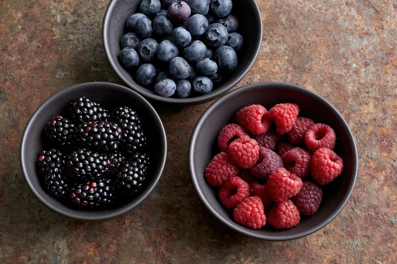 Three dark ceramic bowls each filled with blackberries, raspberries and blueberries arranged on a weathered rust-brown surface