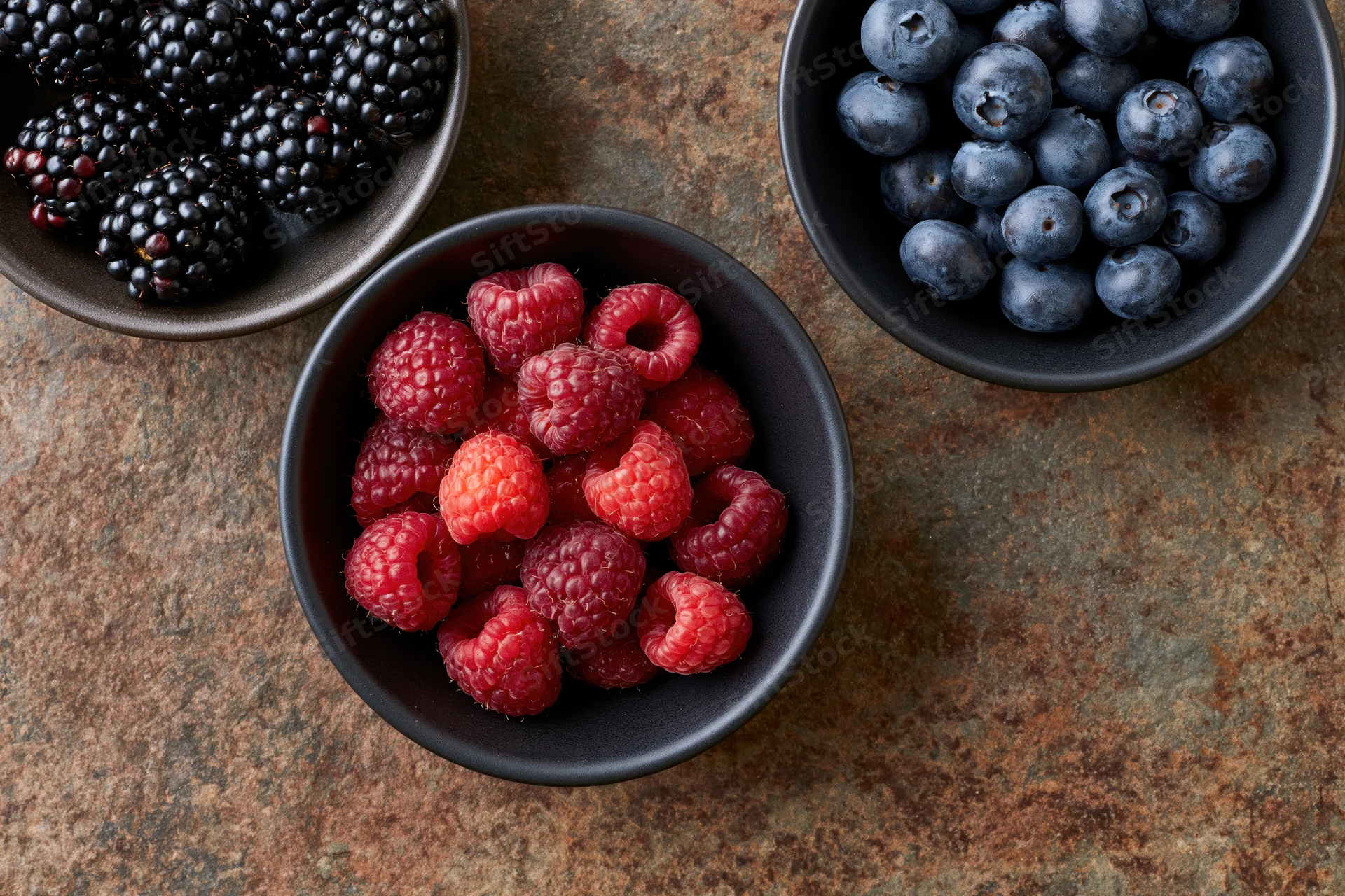 Three dark ceramic bowls filled with raspberries, blueberries, and blackberries arranged on a weathered rust-brown stone surface