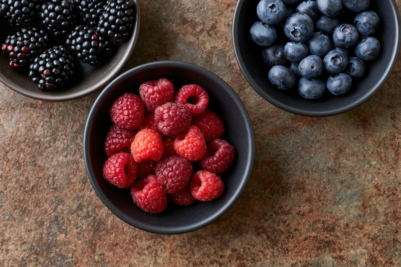 Three dark ceramic bowls filled with raspberries, blueberries, and blackberries arranged on a weathered rust-brown stone surface