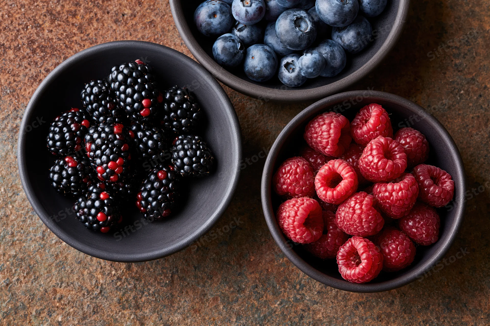 Three dark ceramic bowls filled with blackberries, raspberries and blueberries arranged on a weathered stone surface