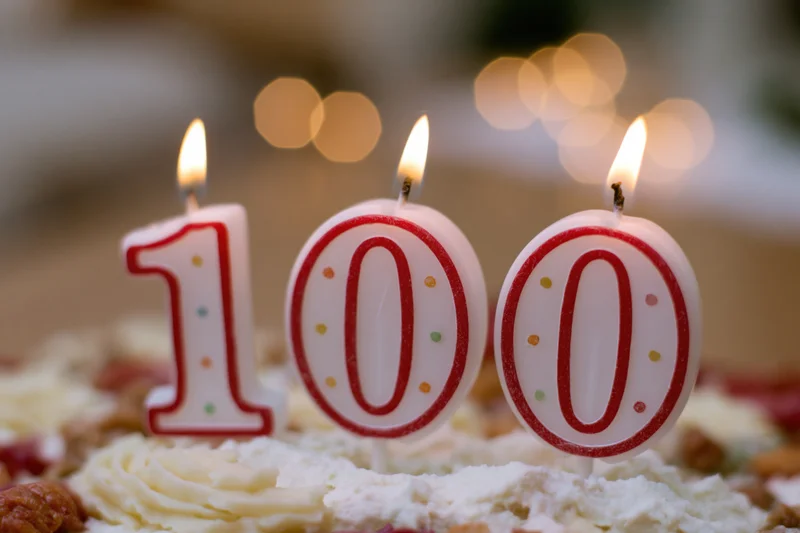 Three lit number candles spelling 100 on a frosted birthday cake