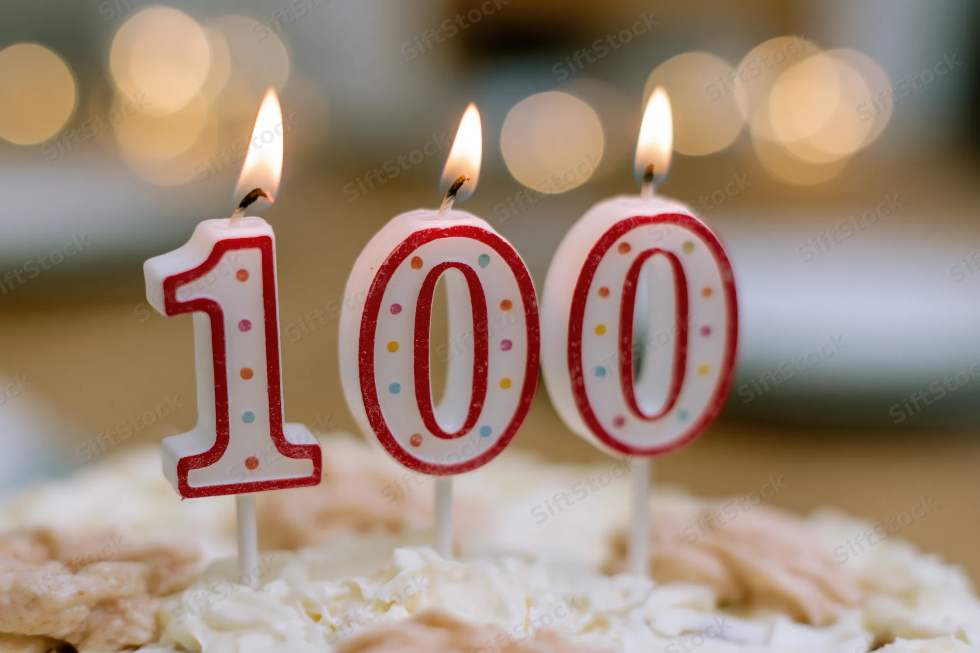 Three lit 100-shaped birthday candles on a frosted cake with blurred lights in the background