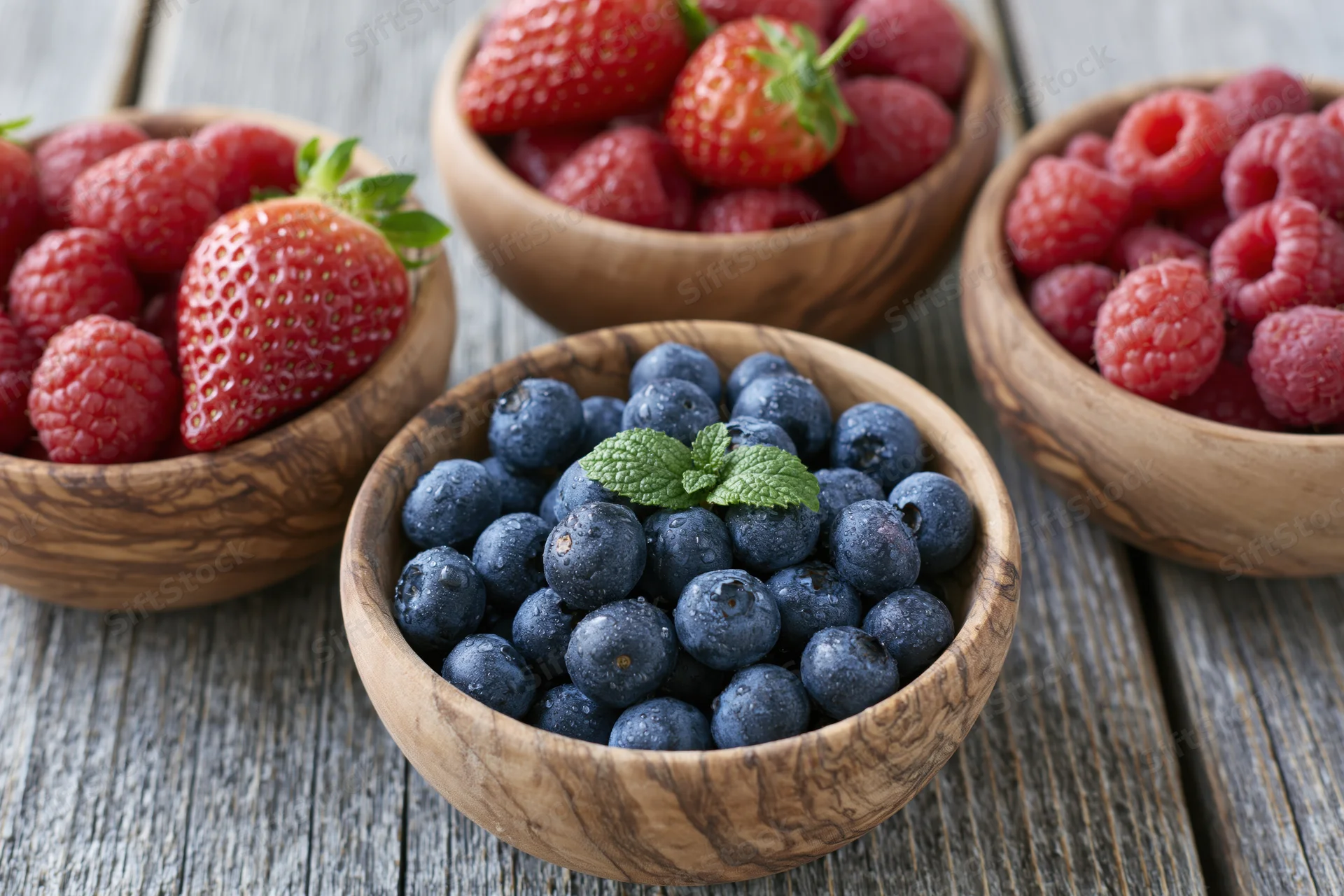 Four wooden bowls filled with blueberries, strawberries, and raspberries arranged on a gray wooden table