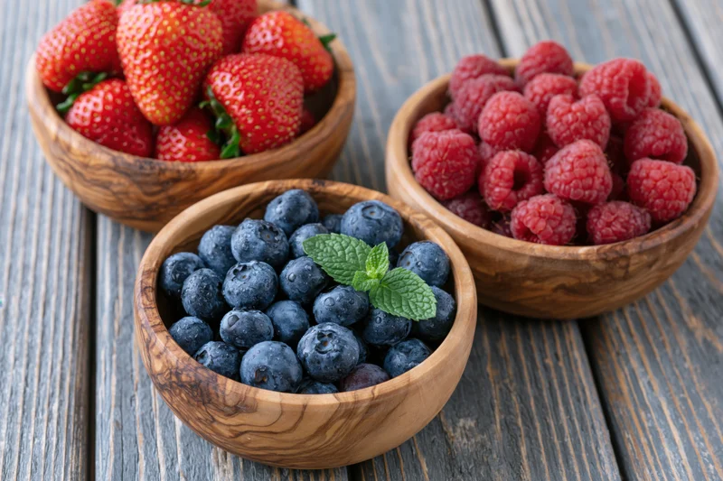 Three wooden bowls filled with blueberries strawberries and raspberries on a gray wooden table