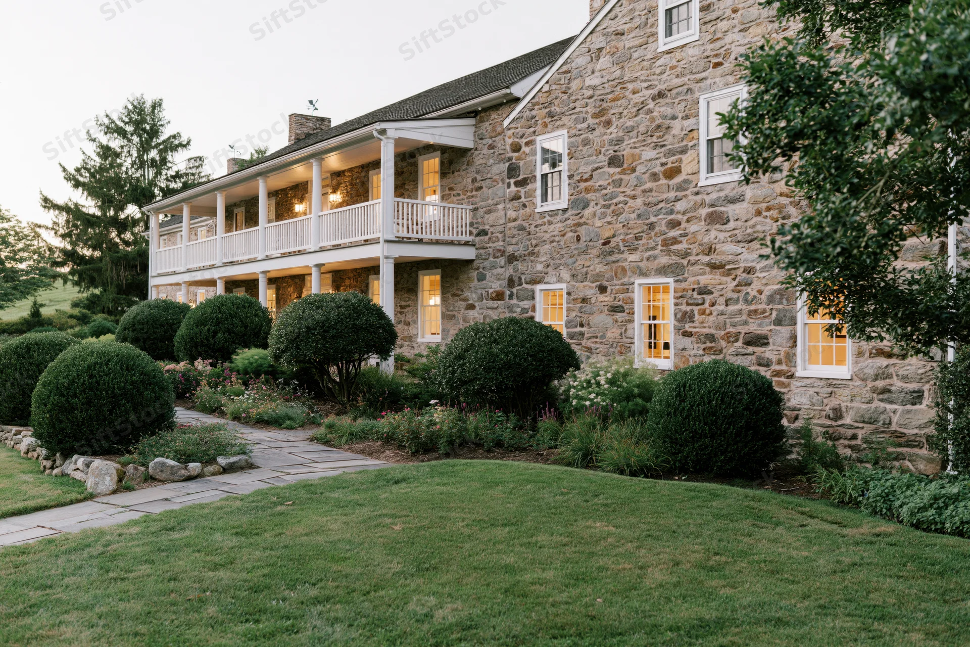 Two-story historic stone farmhouse with white balconies and illuminated windows at twilight, surrounded by manicured lawn and shrubs