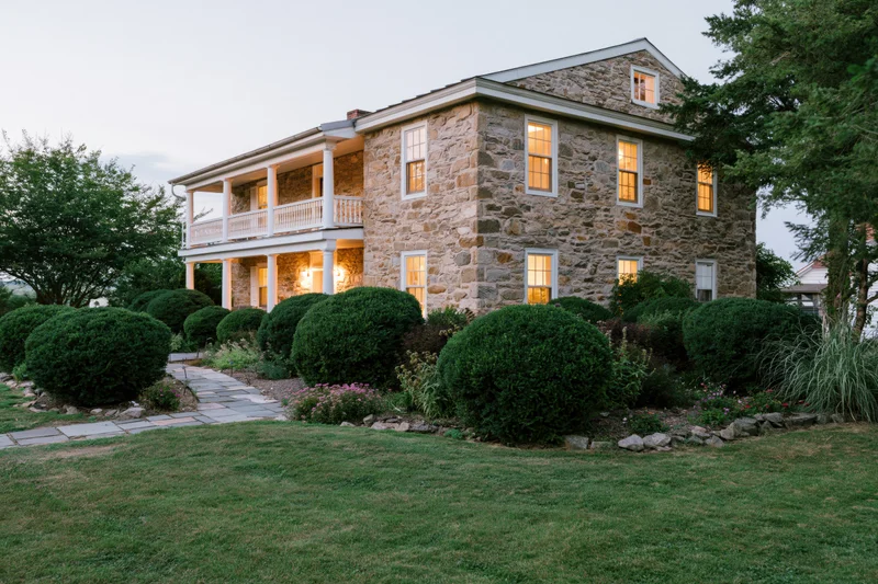 Two-story historic stone farmhouse with white balconies and illuminated windows at dusk surrounded by manicured garden and lawn