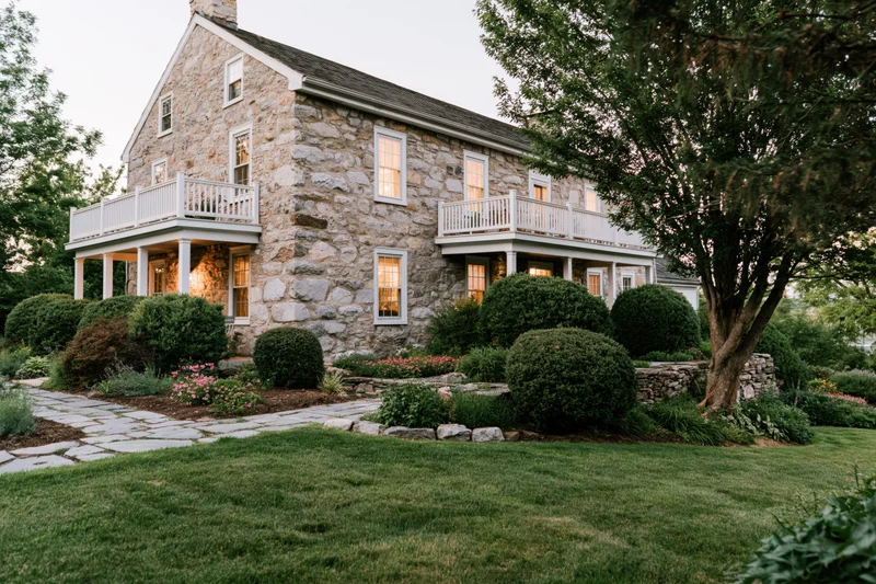 Two-story historic stone farmhouse with white balconies and garden at dusk