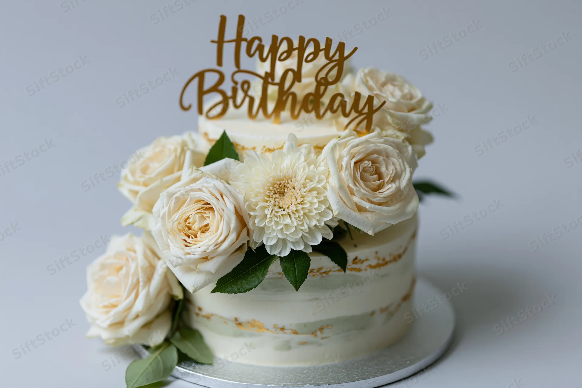 Two-tier white cake decorated with cream roses and a gold happy birthday topper on a silver stand