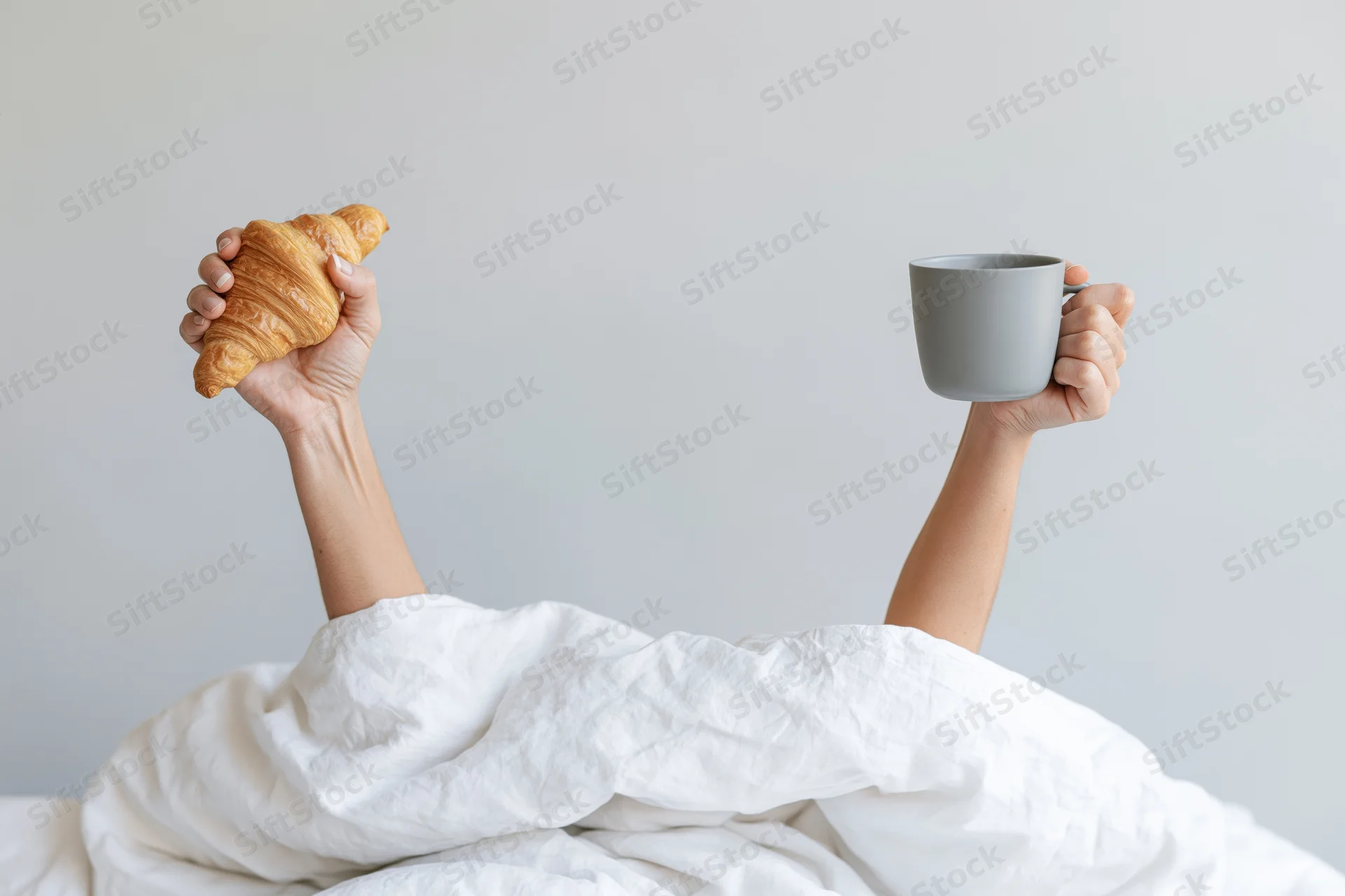 Hands raised from under white bedding holding a croissant and a mug in a bedroom