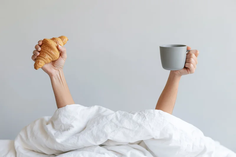 Hands raised from under white bedding holding a croissant and a mug in a bedroom