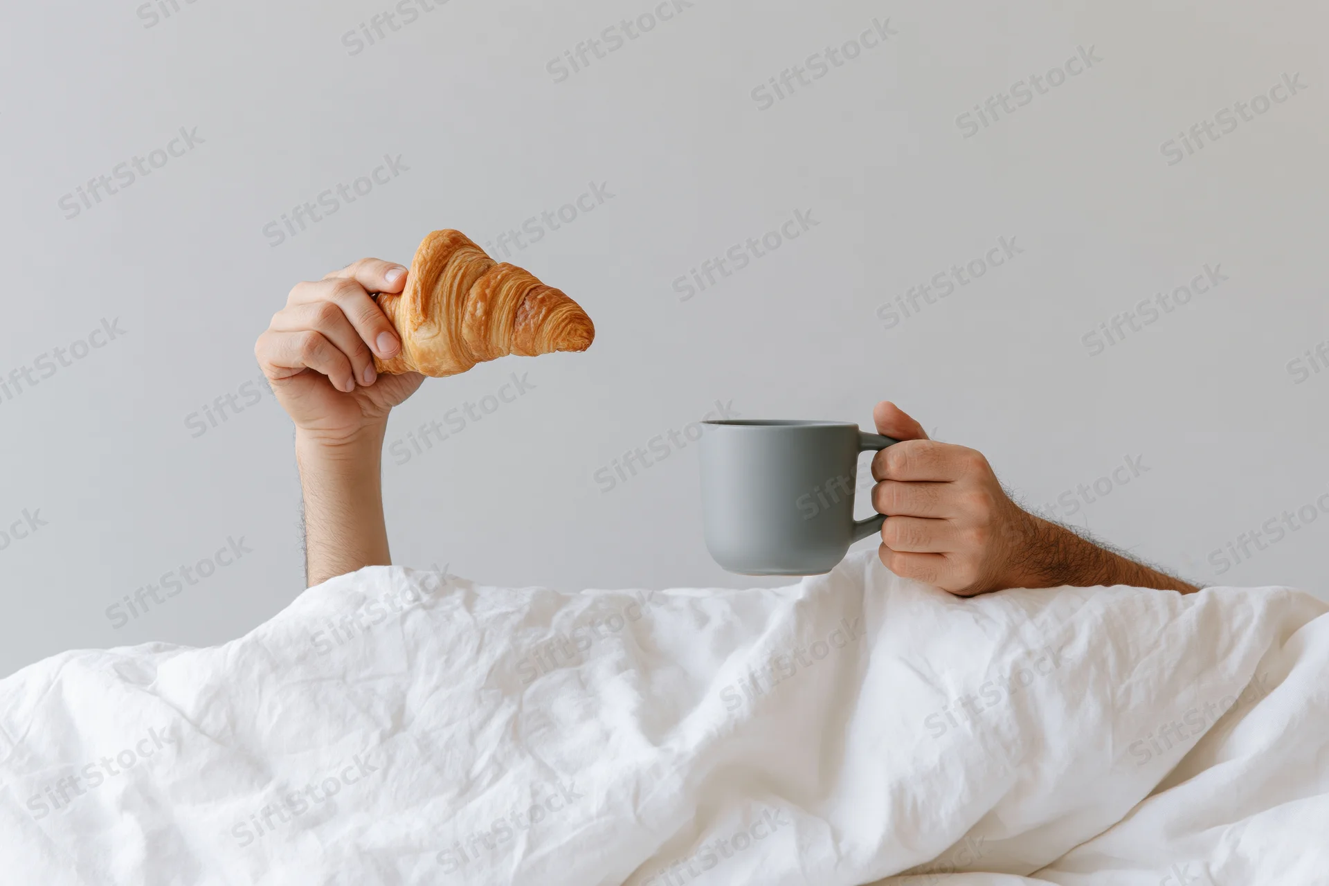 Hands holding croissant and mug emerging from white bedding in bedroom