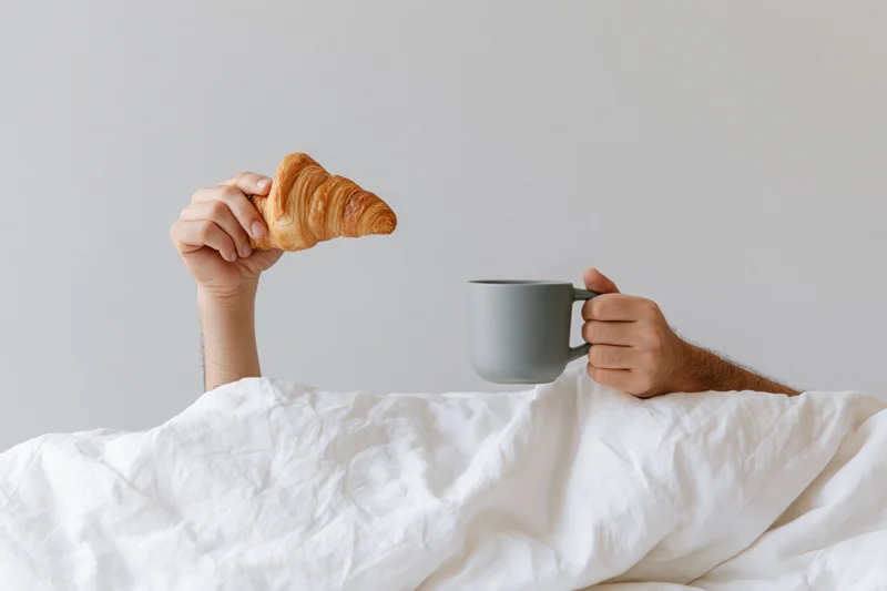 Hands holding croissant and mug emerging from white bedding in bedroom