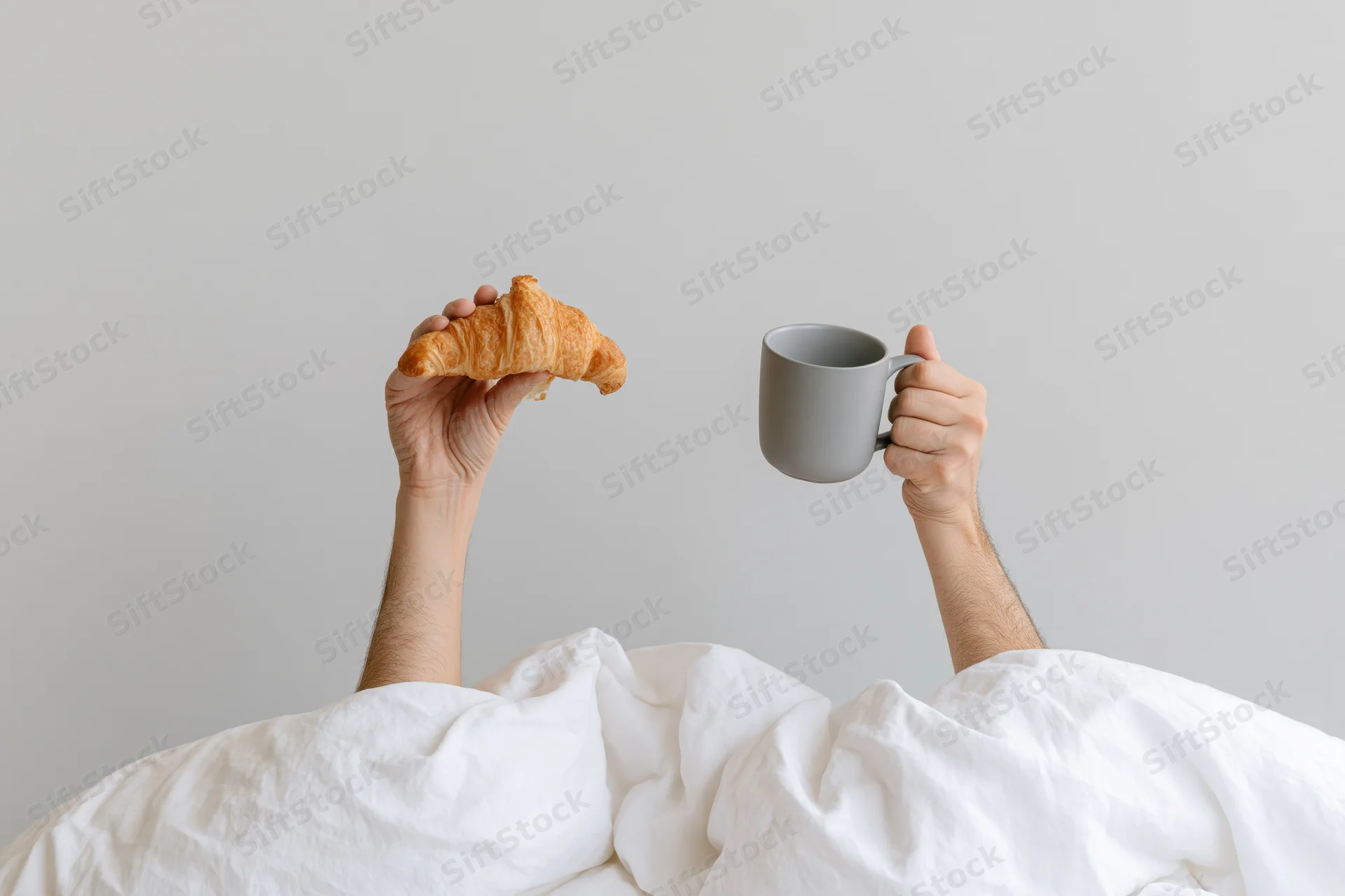 Hands holding croissant and mug raised from under white bedding in morning