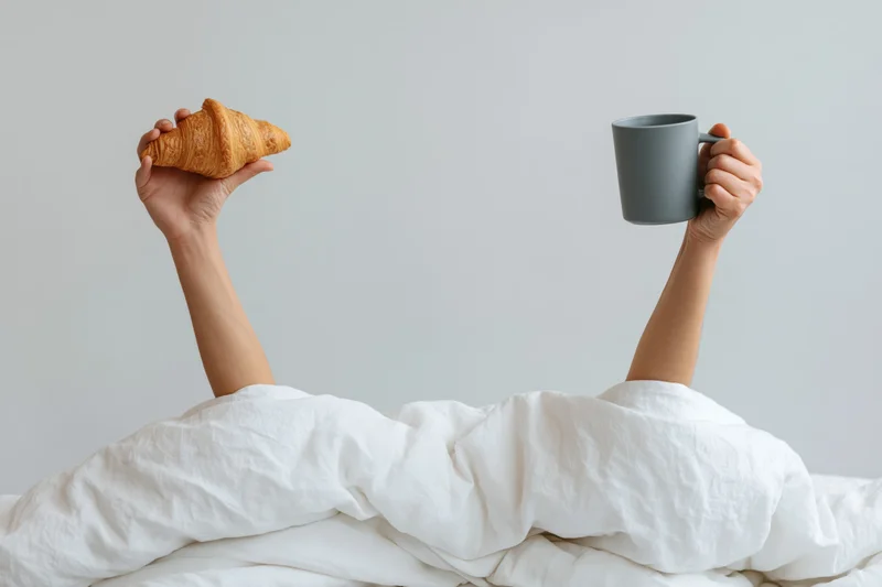 Arms raised from under white bedding holding croissant and mug in morning bed