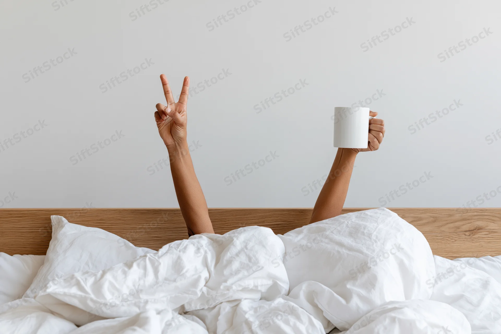 Arms reaching up from under white bedding holding a mug and making a peace sign above a wooden headboard