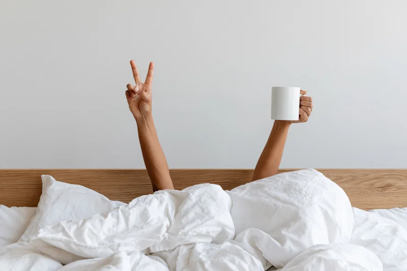 Arms reaching up from under white bedding holding a mug and making a peace sign above a wooden headboard