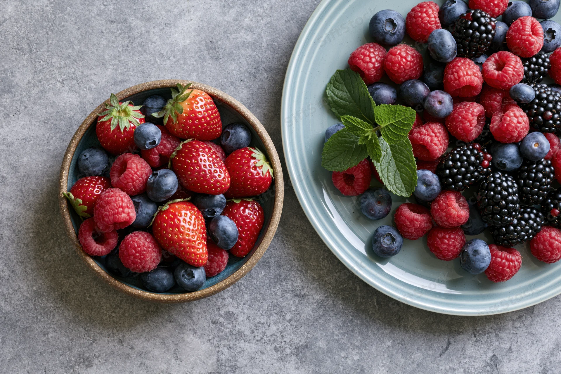 Mixed fresh berries in ceramic bowl and plate on gray stone surface