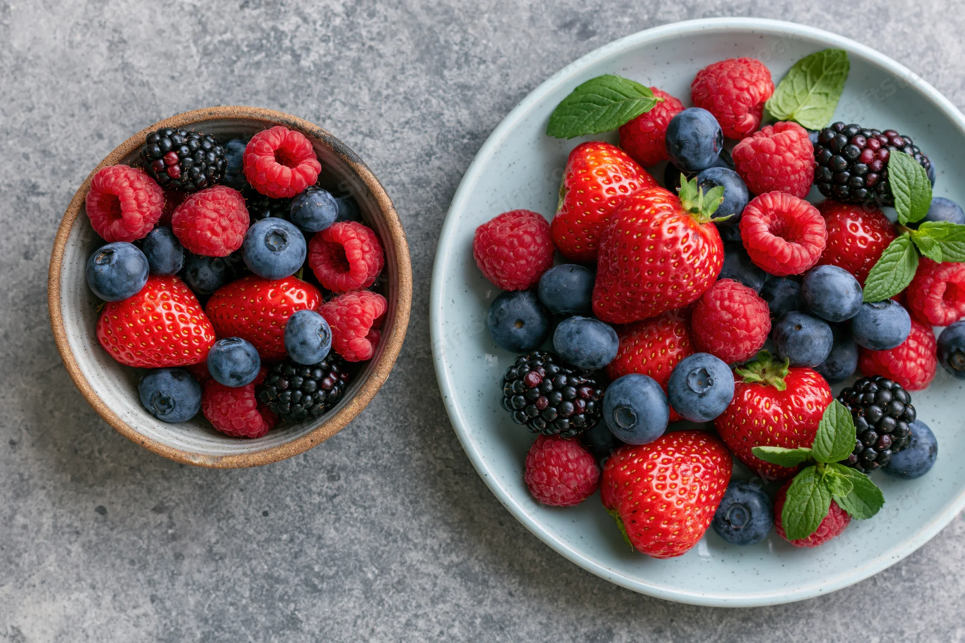 Mixed fresh berries in ceramic bowl and plate on stone surface