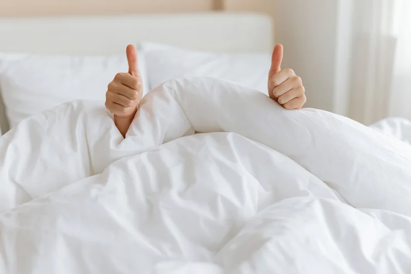 Hands giving thumbs up gesture emerging from under white duvet in bed