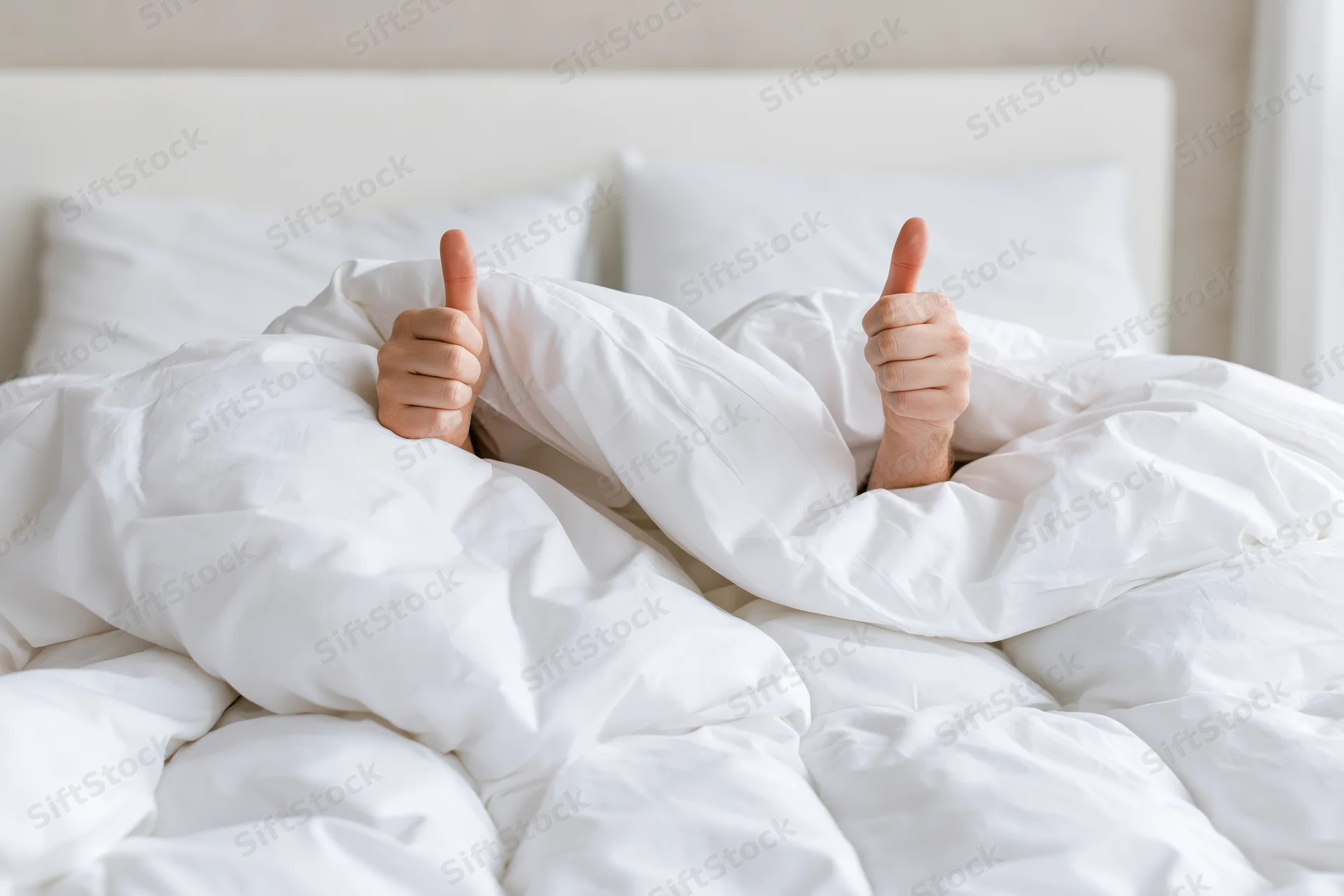 Hands giving thumbs up gesture emerging from under white duvet in bed
