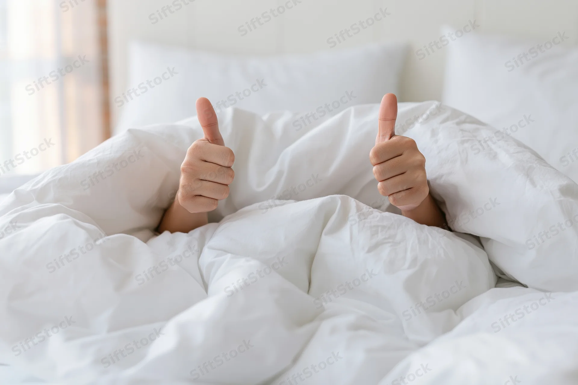 Hands giving thumbs up gesture emerging from under white duvet in bed