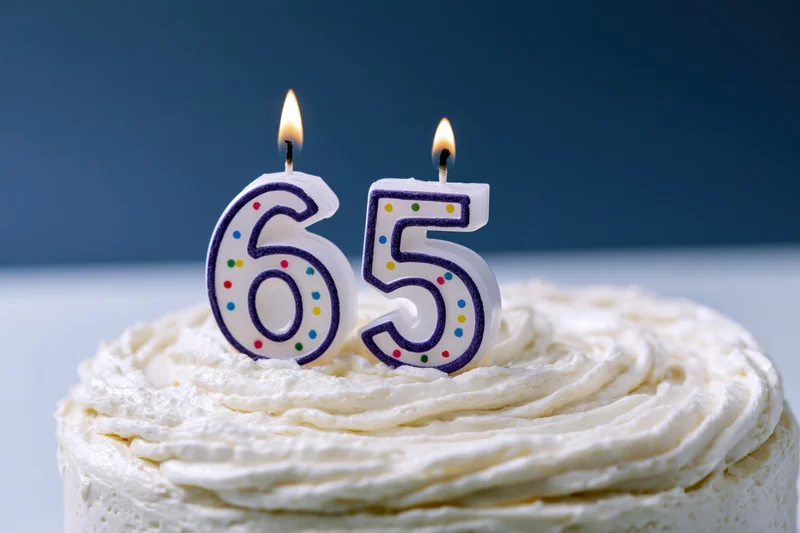 White frosted birthday cake with lit number 65 candles on top