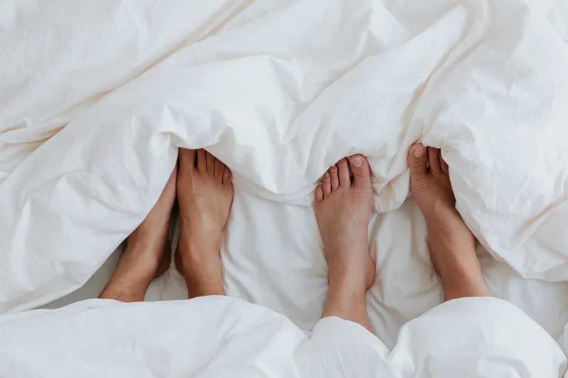 Two pairs of bare feet peeking from under a white duvet in bed