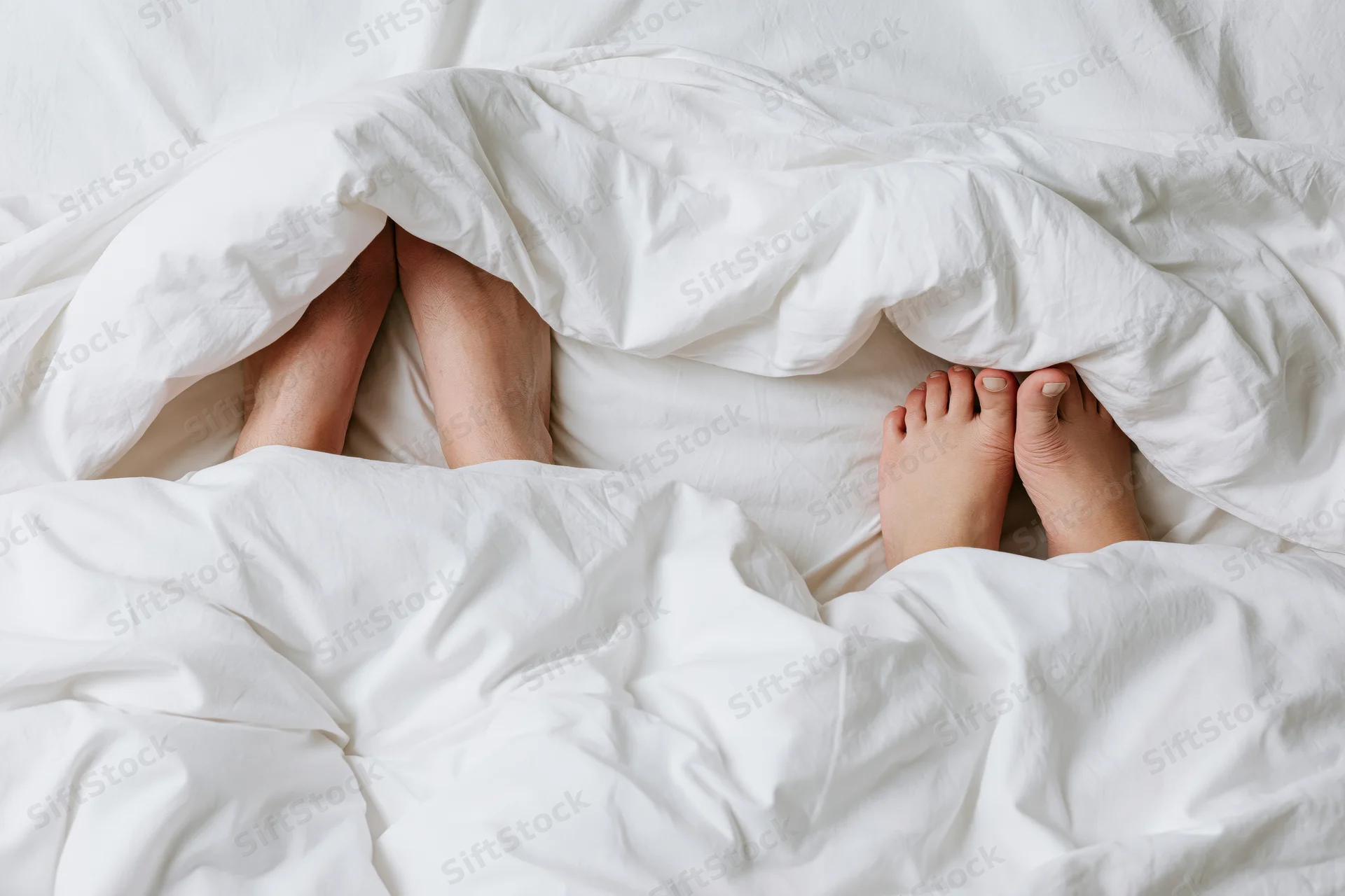 Two pairs of bare feet peeking from under a white duvet in bed