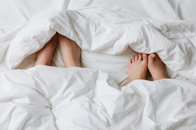 Two pairs of bare feet peeking from under a white duvet in bed