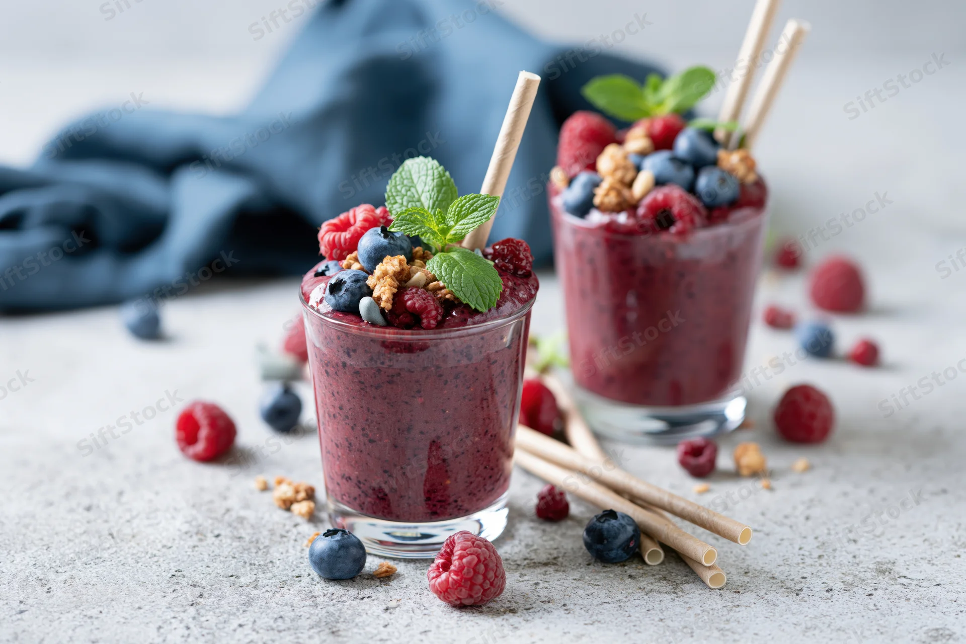 Two glasses of thick mixed berry smoothie topped with fresh berries, mint, and granola on a light countertop