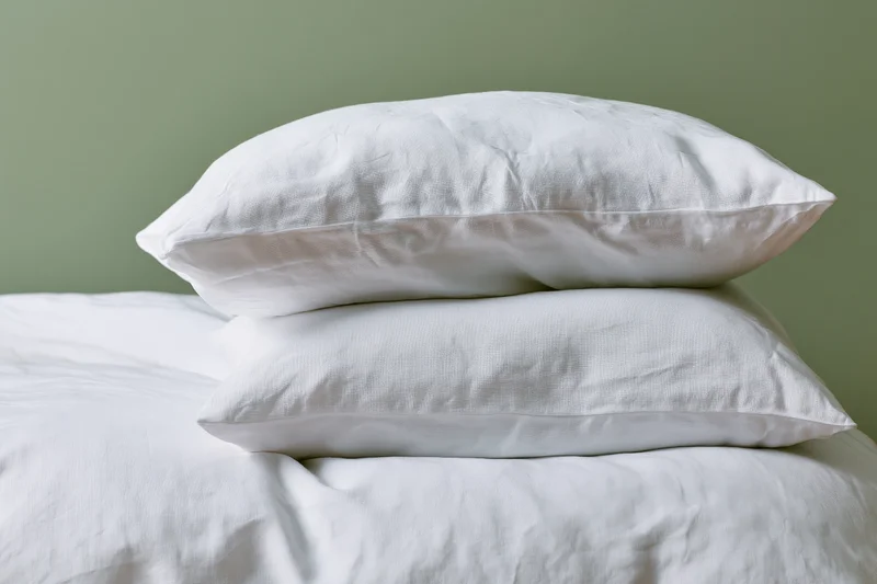 Two white linen pillows stacked on a made bed with white sheets against a green wall