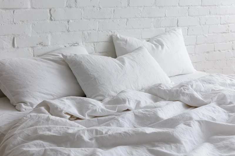 Unmade bed with rumpled white cotton sheets and three pillows against a white brick wall