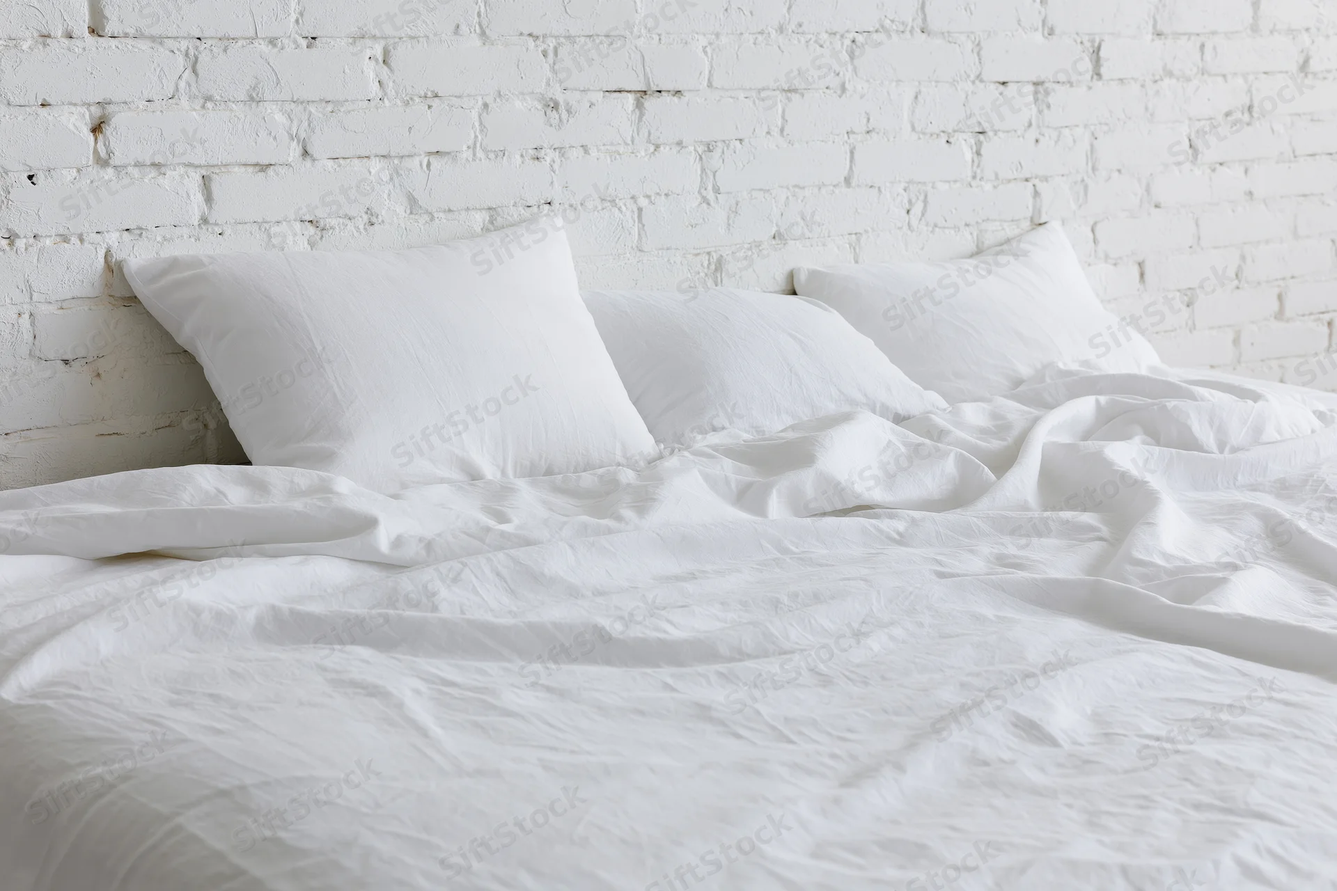 Unmade bed with rumpled white cotton sheets and three pillows against a white brick wall