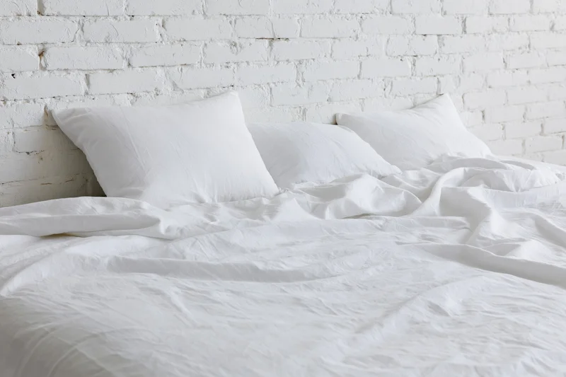 Unmade bed with rumpled white cotton sheets and three pillows against a white brick wall
