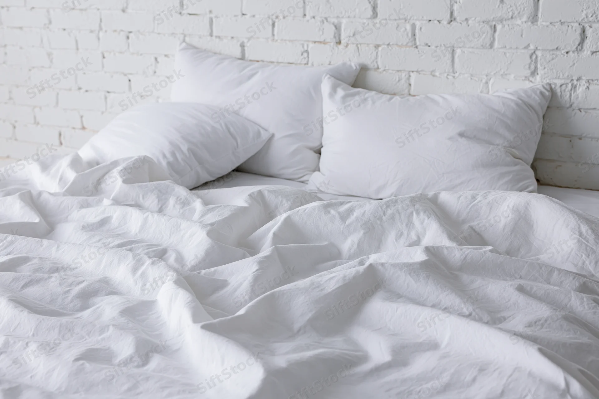 Unmade bed with rumpled white cotton sheets and three pillows against a white painted brick wall