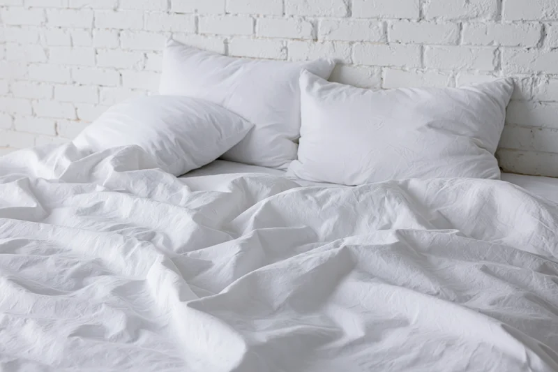 Unmade bed with rumpled white cotton sheets and three pillows against a white painted brick wall