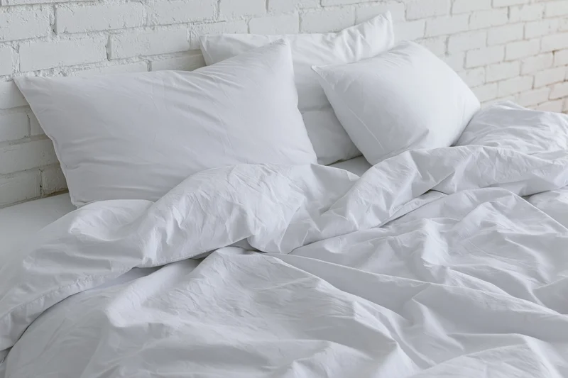 Unmade bed with rumpled white cotton sheets and three pillows against a white brick wall