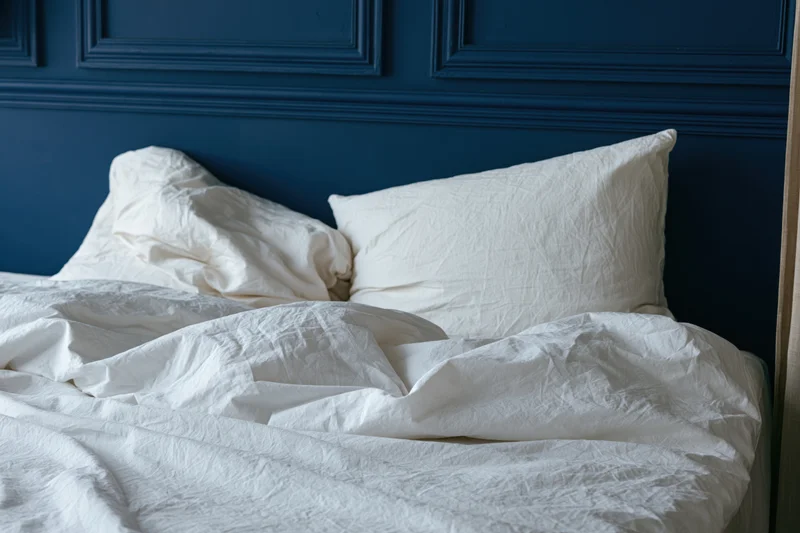 Unmade bed with rumpled white linen sheets and two crumpled pillows against a blue paneled headboard