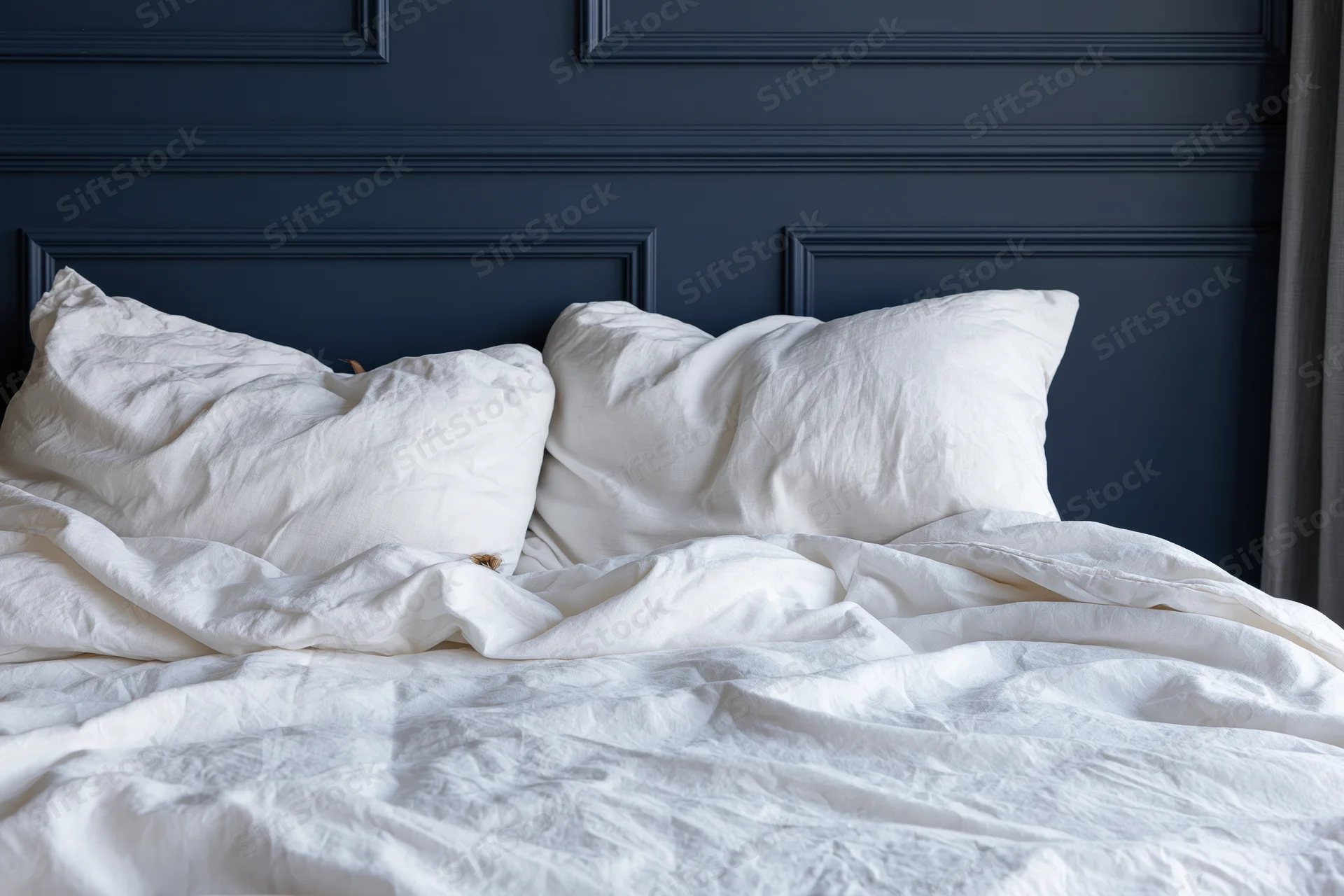 Unmade bed with rumpled white linen sheets and two crumpled pillows against a navy blue paneled headboard