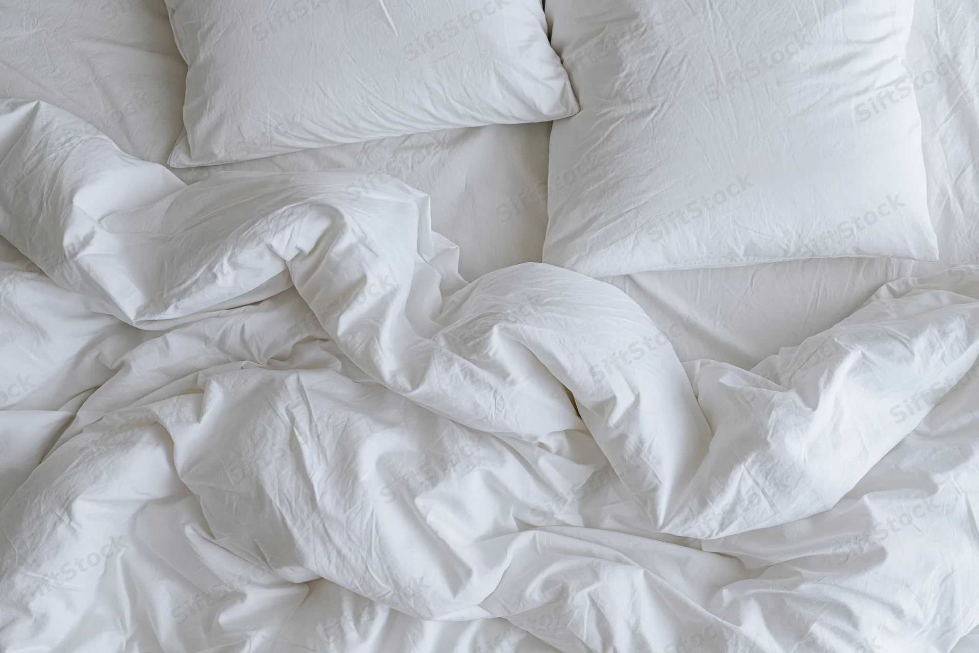 Unmade bed with crumpled white duvet and two flat pillows in bedroom