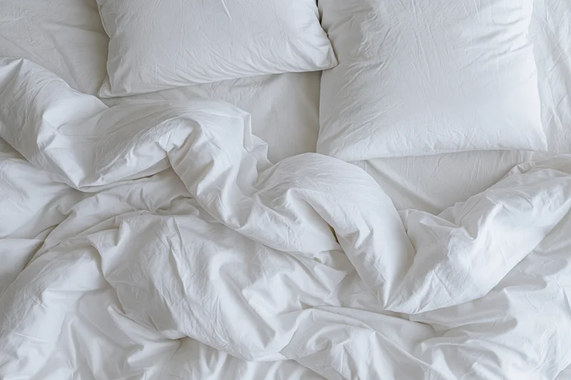 Unmade bed with crumpled white duvet and two flat pillows in bedroom
