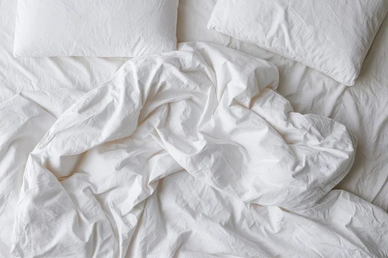 Unmade bed with crumpled white duvet and two flat pillows viewed from above