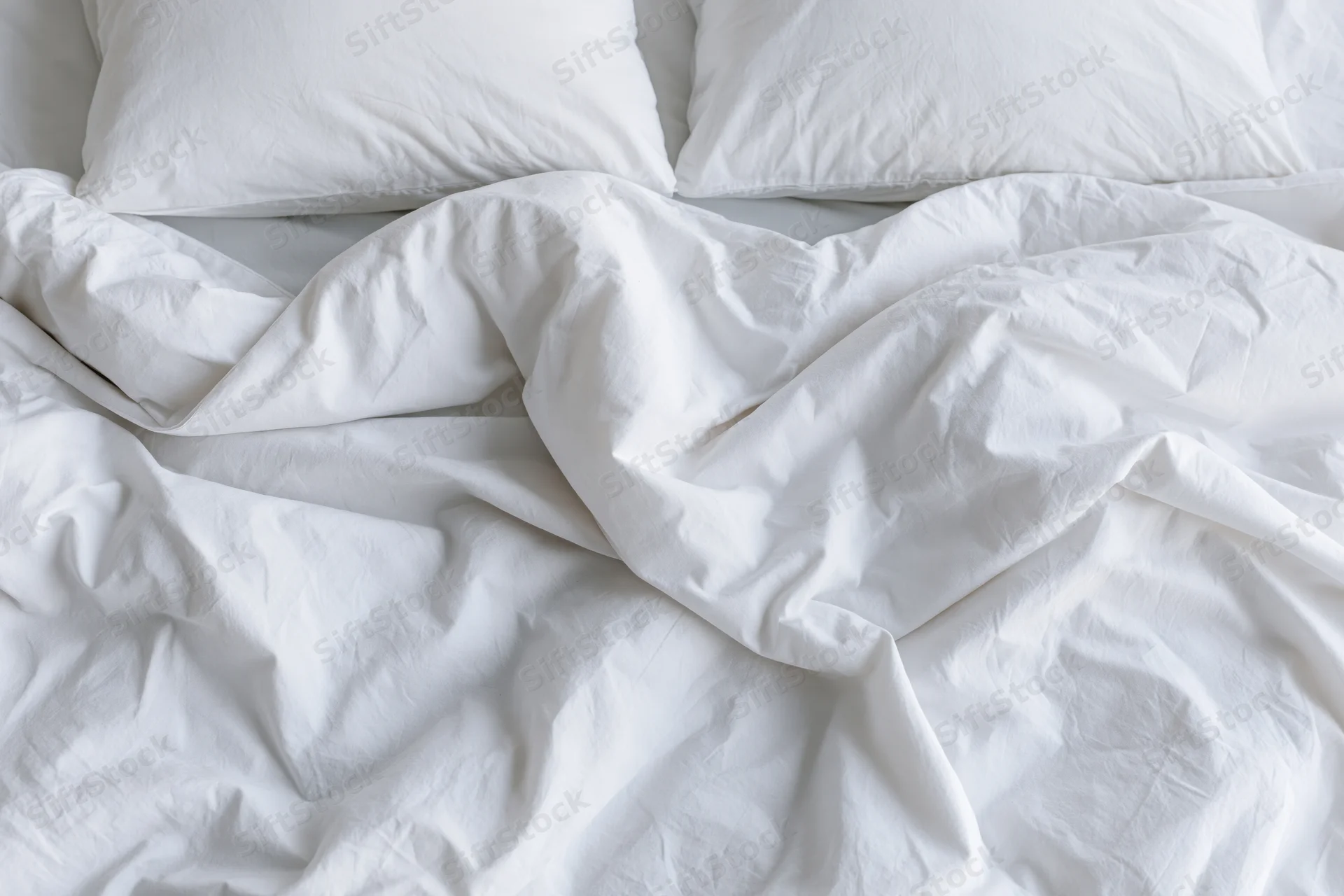 Unmade bed with crumpled white duvet and two flat pillows in bedroom