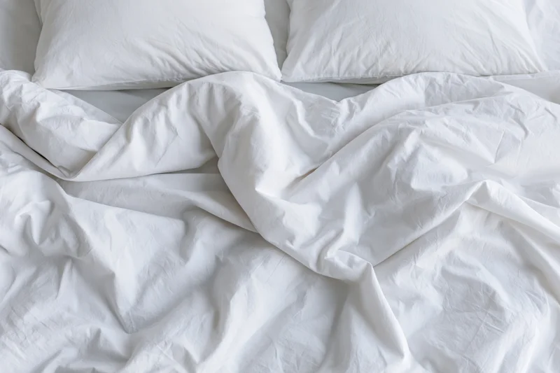 Unmade bed with crumpled white duvet and two flat pillows in bedroom
