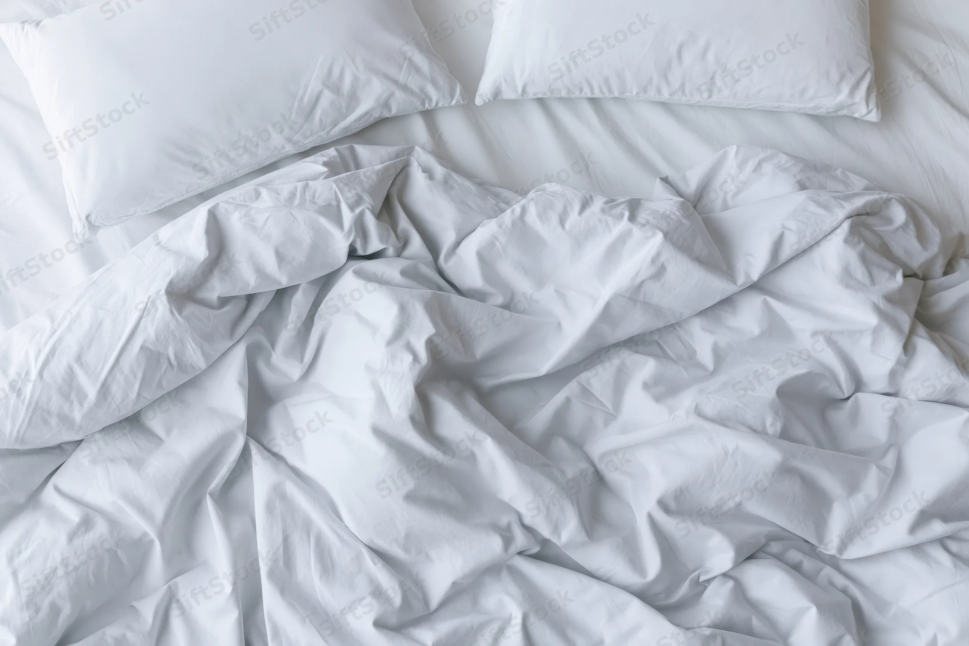 Unmade bed with crumpled white duvet and two flat pillows viewed from above
