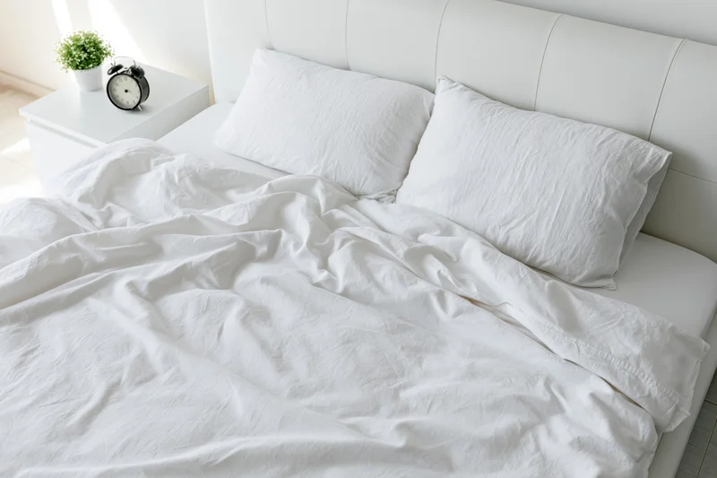 Unmade bed with white rumpled cotton sheets and two pillows in a bright minimalist bedroom
