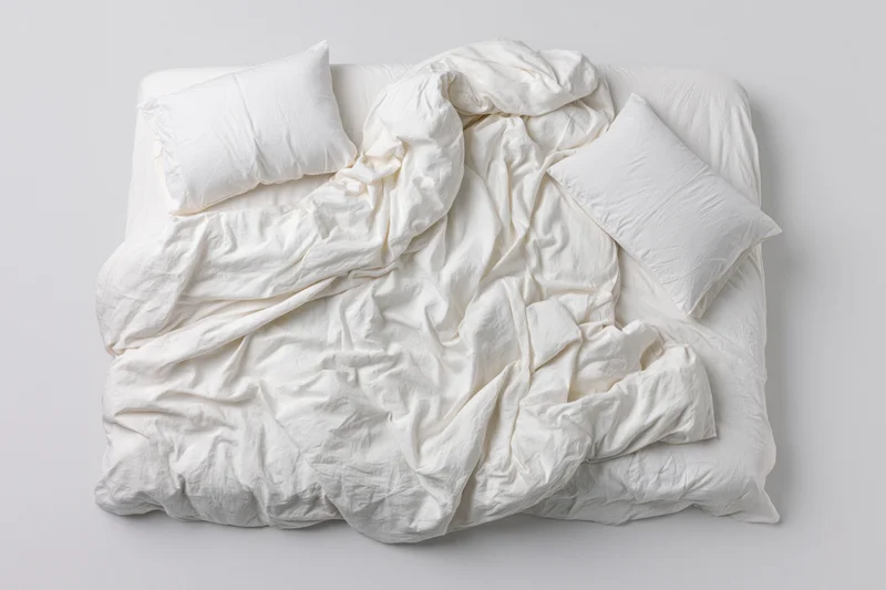 Unmade double bed with rumpled white cotton sheets and duvet on plain background