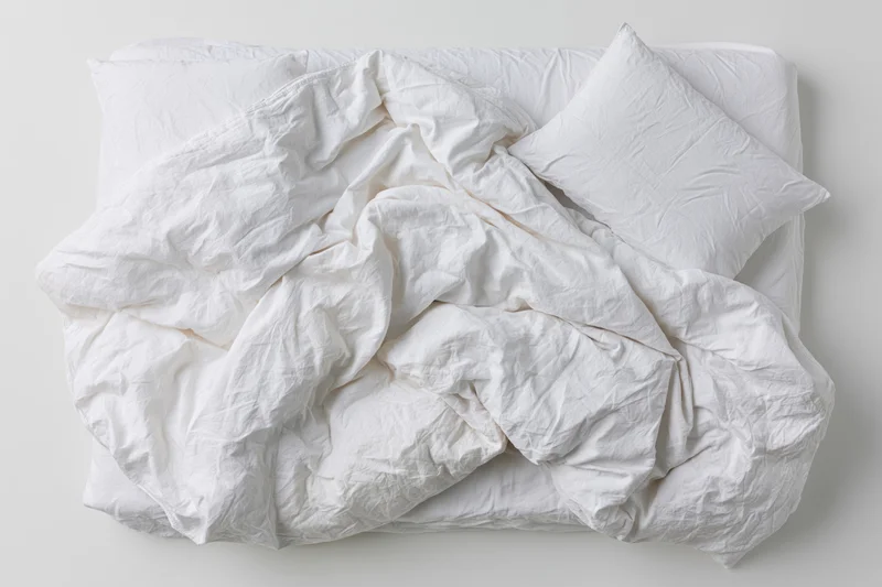 Unmade double bed with rumpled white cotton sheets and duvet on a plain surface